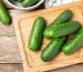 A wooden cutting board with several cucumbers placed on it additional cucumbers in a bowl in the background table surface underneath