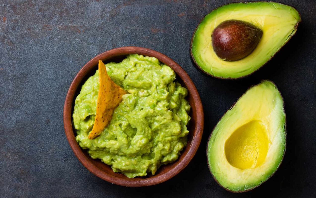 A bowl of guacamole with a tortilla chip accompanied by two avocado halves on a dark background