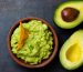 A bowl of guacamole with a tortilla chip accompanied by two avocado halves on a dark background