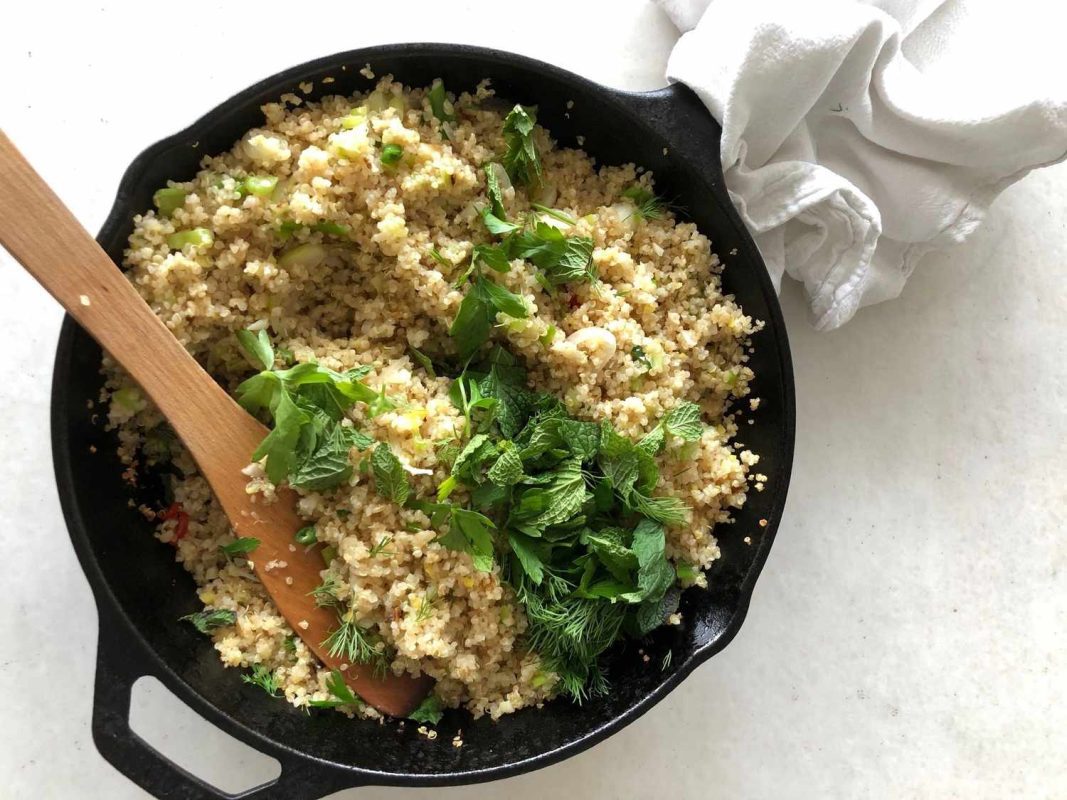 A skillet containing cooked quinoa garnished with fresh herbs and a wooden spoon placed within